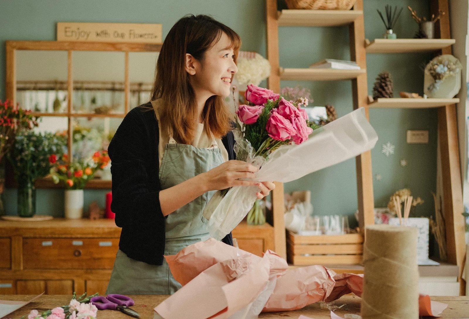 Optimistic Asian female florist wearing apron making flower bouquet with roses while standing in floral workshop